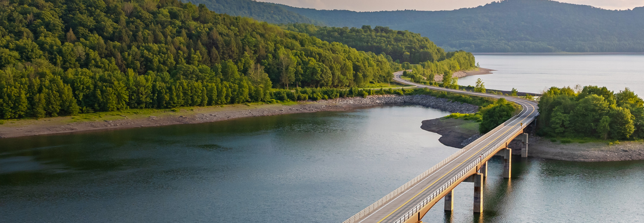 A highway road elevated over a lake and winding out toward a lush green mountain range