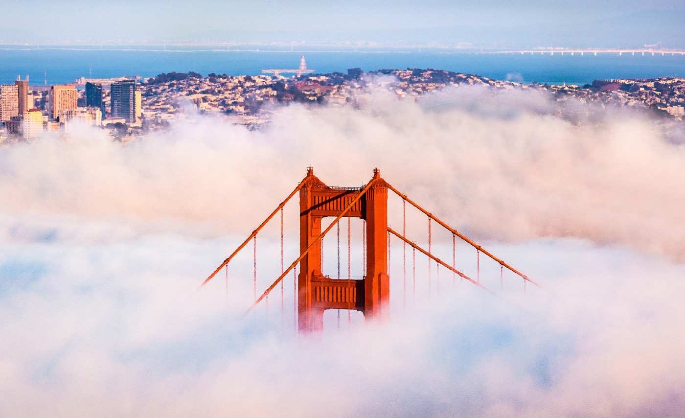 The Golden Gate Bridge poking out from a vast foreground of clouds with the city scape in the far background.